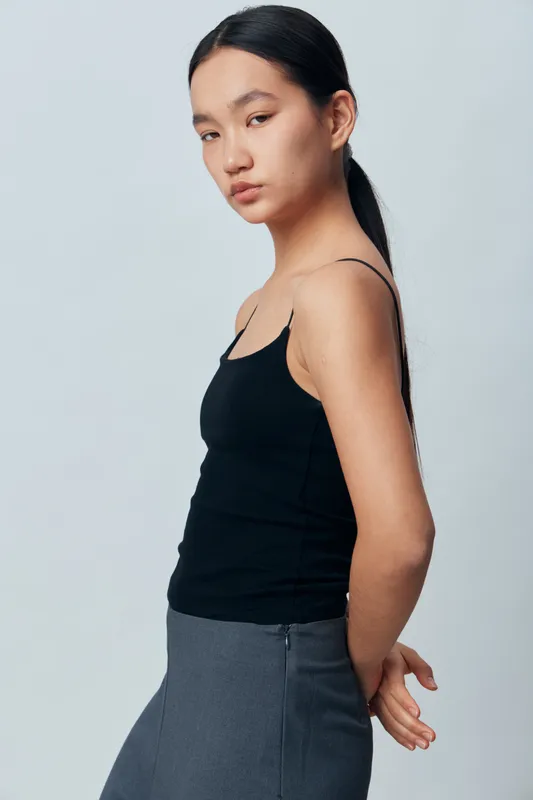Young woman with long black hair wearing a black tank top and gray skirt standing in profile with hands behind back against plain light background.