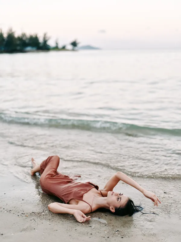 Solitary figure lies on wet sand near calm sea, sunlight soft, relaxed pose, flowing dress, gentle waves, tranquil coastal moment captured in warm pastel hues.