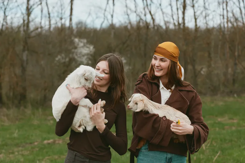 Goat goats kid animal animals lifestyle outdoor countryside women sharing a lighthearted moment with two kids held by friends in an open field, soft daylight, casual outfits, warm smiles,