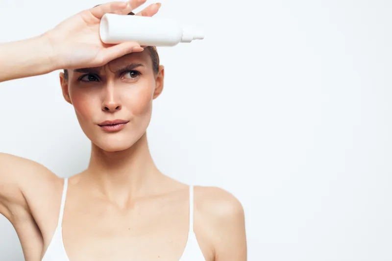 Young woman with clear skin holding white cosmetic bottle on forehead. Studio shot on white background. Concept of skincare, beauty, health, and wellness. Soft natural lighting.