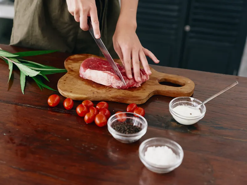 Женщина с ножом в руке нарезает свежий стейк из мяса для жарки на кухне