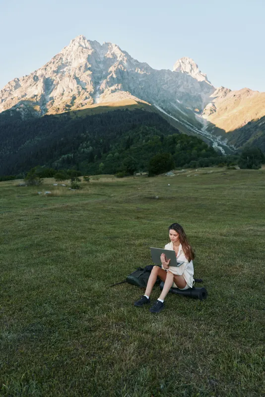 A student sits on a grassy meadow with a laptop in a scenic mountain landscape. She concentrates on her screen, exuding calm focus and a sense of productive solitude.