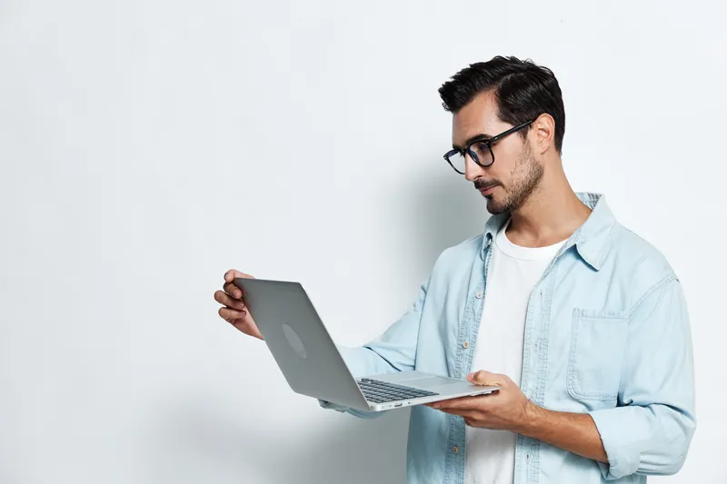 Thoughtful young man wearing glasses, holding a laptop, looking at the screen, isolated on a light solid background. Technology lifestyle concept.