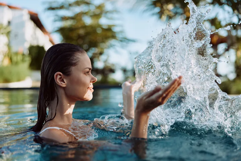 Young woman in a pool splashes water with her hands, bright sunlight, carefree mood and a refreshing summer vibe captured in a candid moment