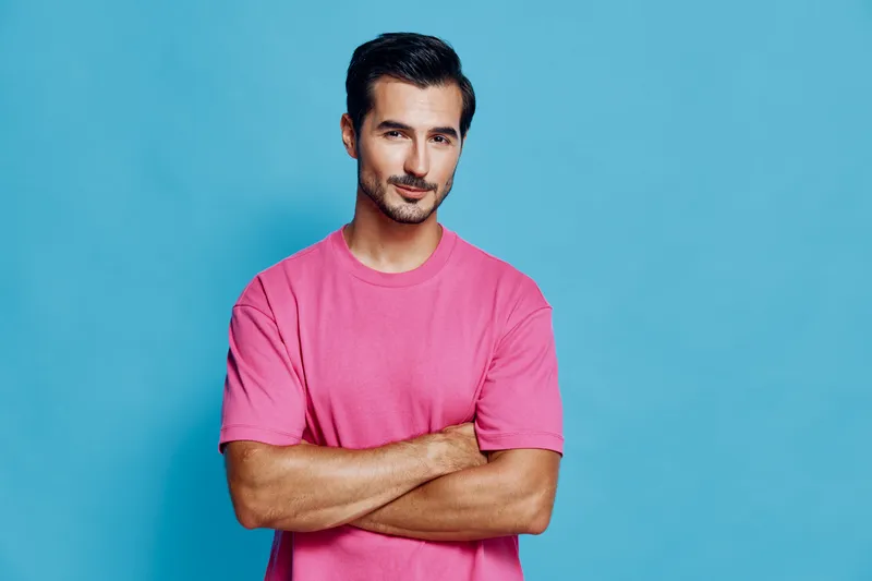 Confident young man with crossed arms wearing a casual pink t-shirt stands against a bright blue background, smiling at the camera. Vibrant lifestyle concept.