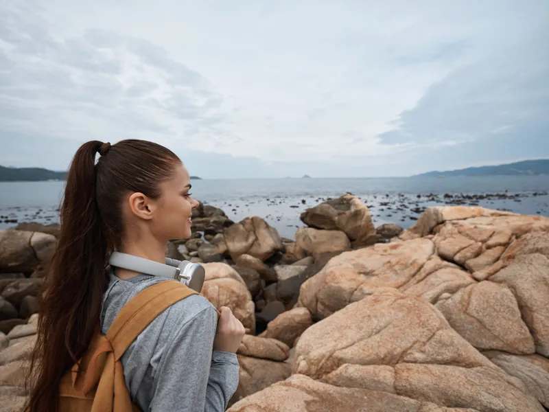 A young woman of Asian descent wearing casual clothing with a backpack, gazing contemplatively at the seaside landscape, evoking feelings of adventure and exploration