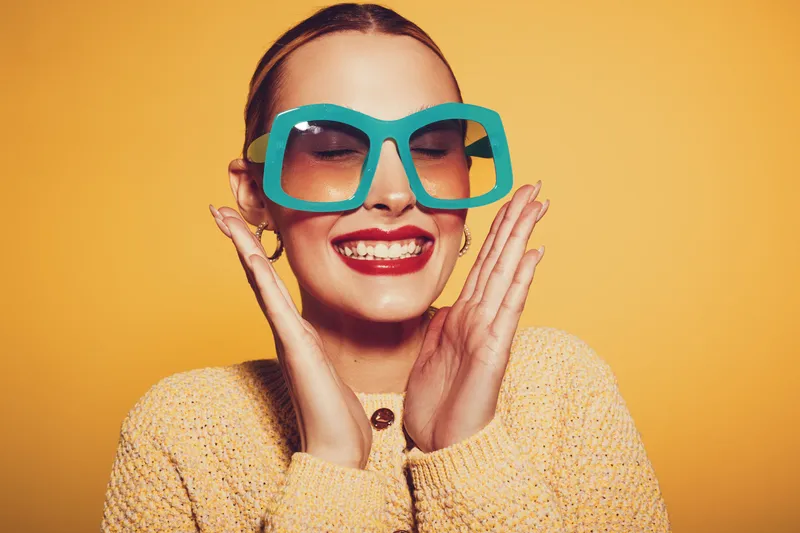 Smiling woman with large turquoise glasses and red lipstick wears yellow sweater, posing with hands near face on bright yellow background. Concept of fashion, joy, and vibrant style.
