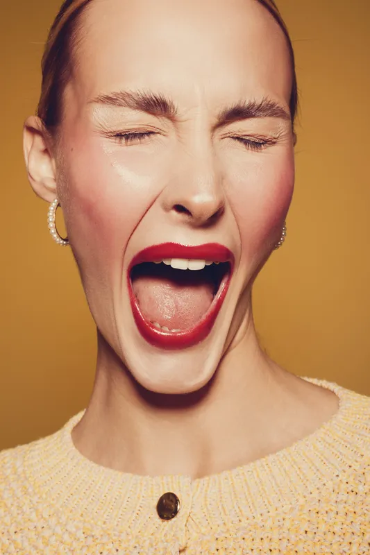 Close-up of a young woman with red lipstick and closed eyes shouting or singing against a warm yellow background. Concept of emotion, expression, and beauty portrait.