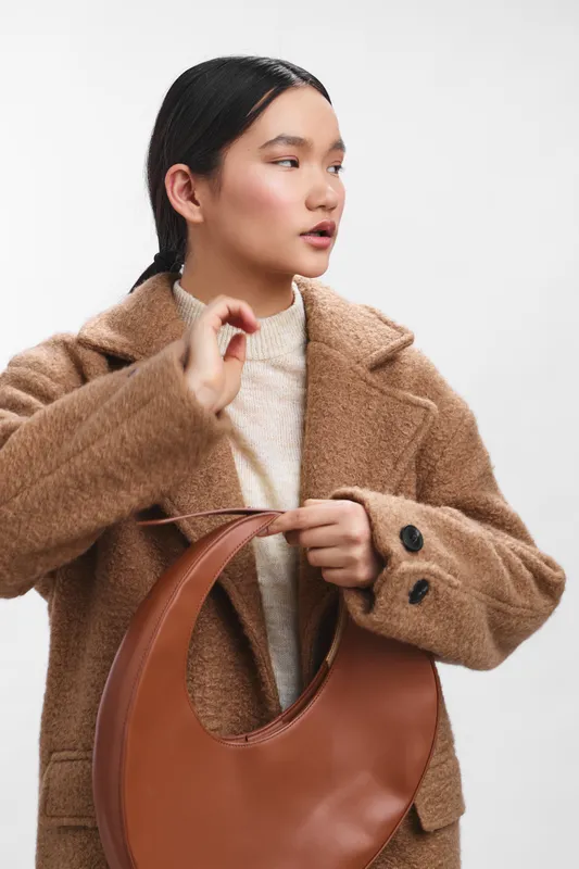 Asian woman wearing a tan wool coat holds a rounded leather bag in a clean studio setting. The minimalist fashion shot highlights texture, style, and confidence, creating a contemporary and polished