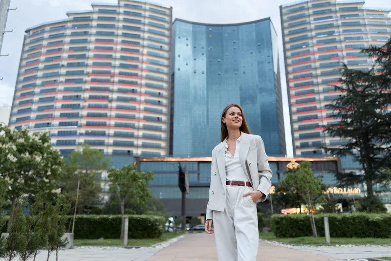 business woman in beige suit walking confidently in modern cityscape with office towers and green park background