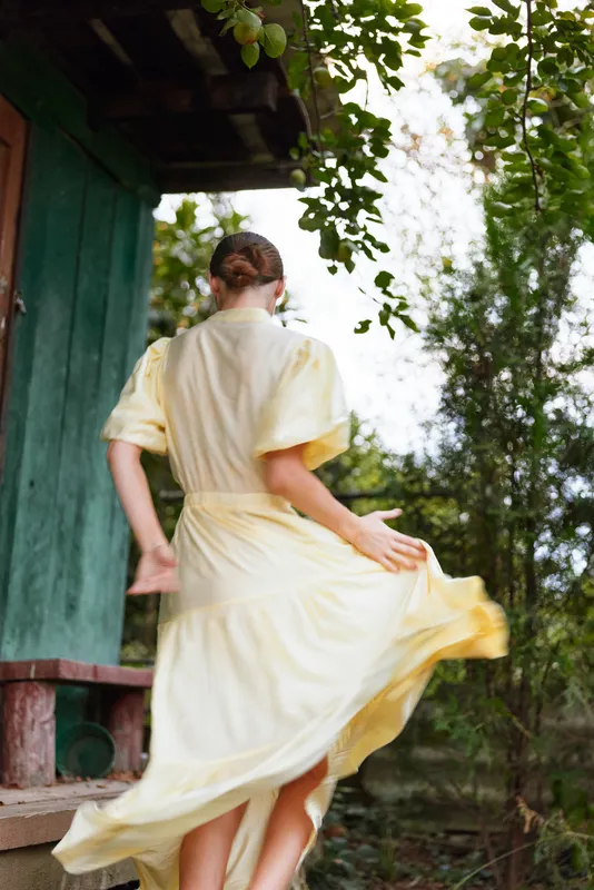 Woman in a flowing yellow dress walks away from the camera in a leafy garden, wind catching the hem, creating a sense of motion and sunny summer style in a natural setting with greenery and a rustic