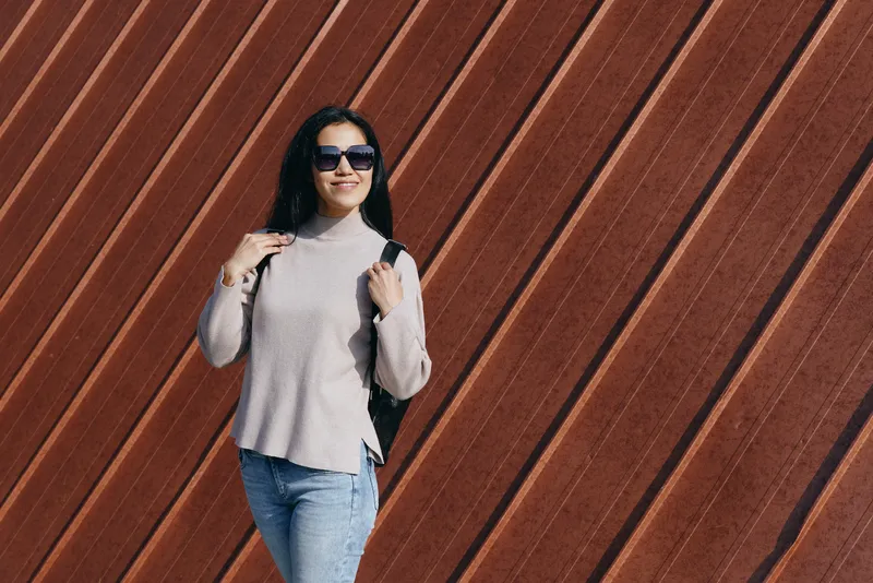 Woman with backpack and sunglasses posing against a diagonal wooden wall, casual fashion, urban style, outdoor scene, lifestyle image capturing confident streetwear look and relaxed demeanor