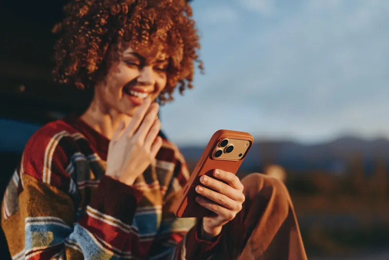 A woman lifestyle outdoors portrait, curly hair smiling female holds a phone and touches her face for communication, call chat on screen, evening sunset mountains in background, relaxed mood
