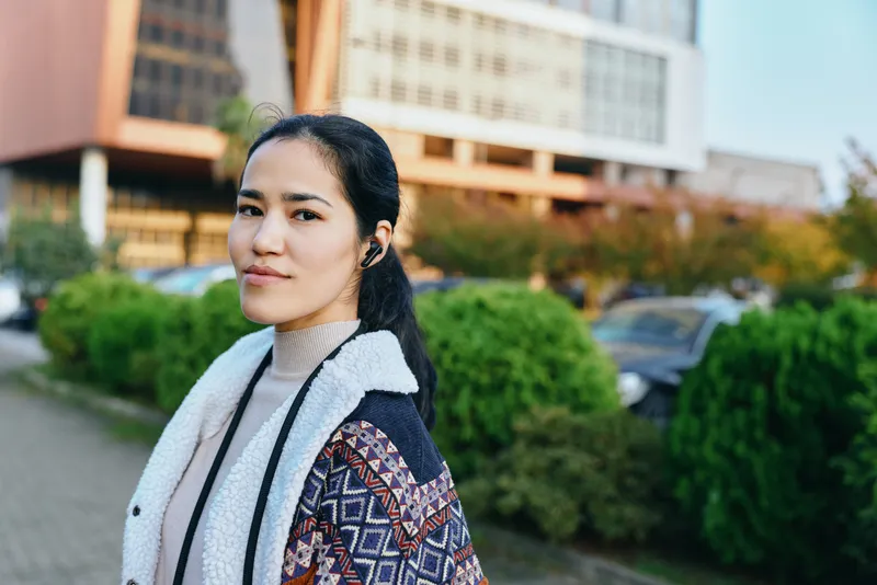 Young woman outdoors wearing a patterned cardigan and neutral scarf, standing beside greenery with urban buildings in the background, casual street fashion and everyday style.