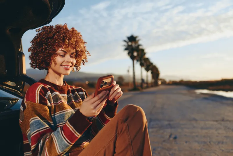 Woman lifestyle smartphone outdoors sunset curly hair smiling casual sweater, young woman sits by car trunk, looking at phone, relaxed posture, golden hour light, palm lined road