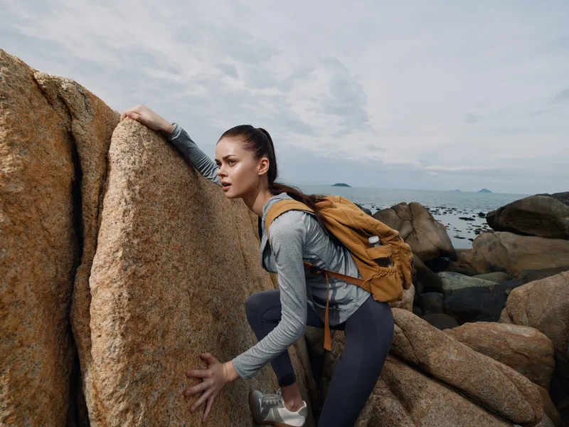 Active young woman climbing rocky terrain, wearing athletic gear and a backpack, demonstrating determination in a coastal landscape