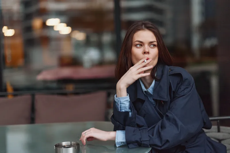 Young woman with long brown hair wearing a dark trench coat sits at a glass table outdoors while thoughtfully looking to the side and holding a cigarette near her mouth.