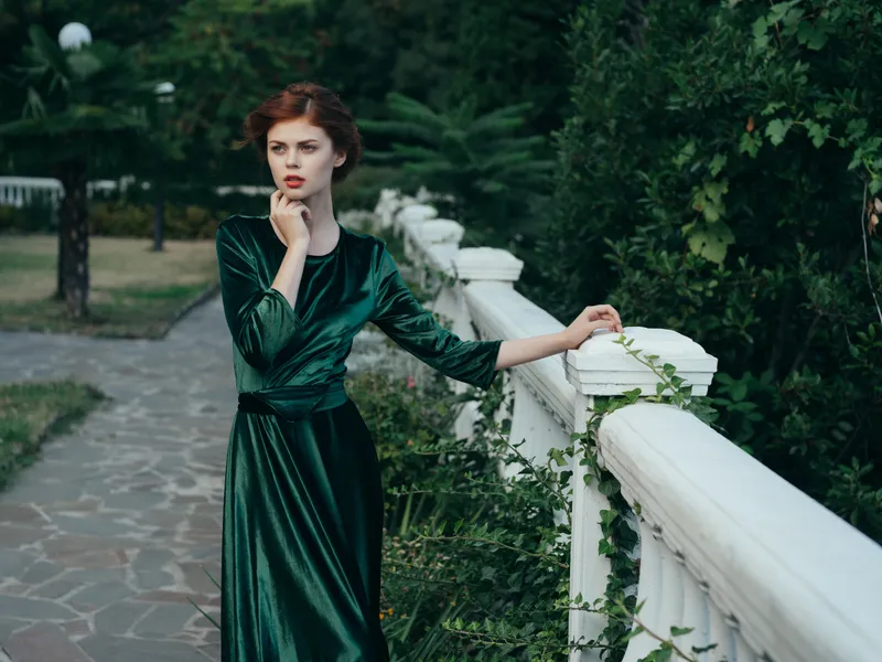 Женщина в зеленом платье гуляет по природному парку, опираясь на перила