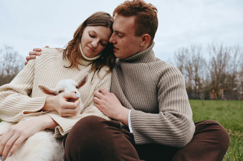 Couple outdoors in cozy sweaters cuddle a gentle goat kid on a grassy field, sharing affection and calm happiness, a candid lifestyle moment with a rustic, natural backdrop.