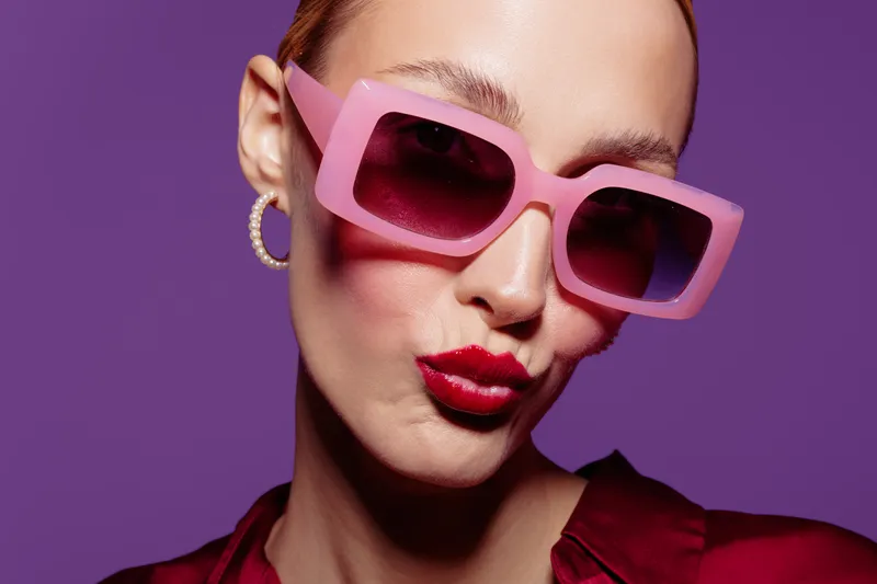 Close-up of a woman wearing pink sunglasses and red lipstick posing against a purple background. Fashion portrait with bold makeup and stylish accessories in studio lighting.