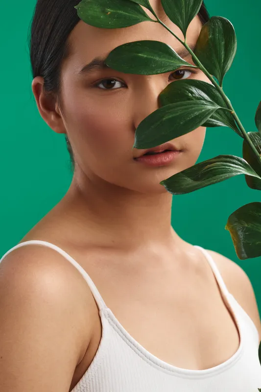 asian woman gazes softly in a close up portrait with a green backdrop and leafy foreground, conveying calm beauty, natural style, and a contemplative mood.