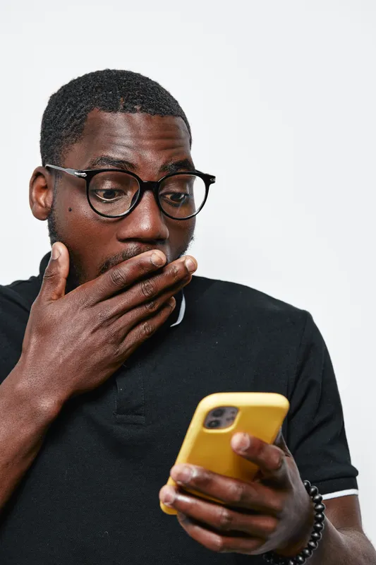 Black man with dark skin and glasses looking surprised at smartphone screen, wearing black polo shirt and bracelet, isolated on white background with emotional reaction.
