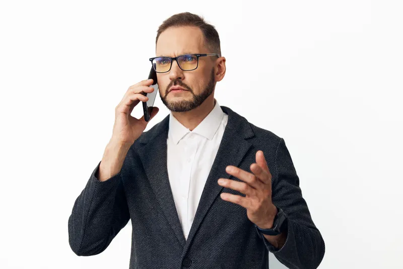 A professional man on a solid colored background speaks on a mobile phone, displaying focus, confidence, and determination in a modern business setting.
