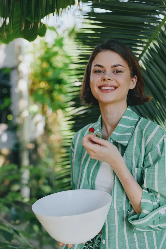Woman smiling outdoors holds a white bowl, wearing a green striped shirt, sunlit garden backdrop. Natural candid moment capturing joy, style, and calm confidence in everyday life.