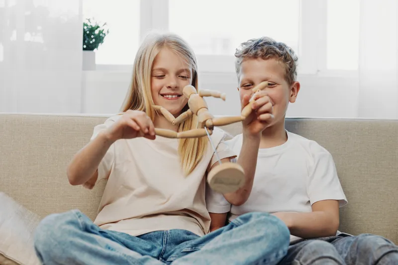 Two happy children sitting on sofa playing with wooden mannequin toy, smiling and enjoying playful moment in bright living room interior.