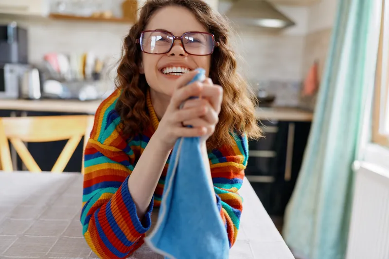 A cheerful young woman in a colorful striped sweater cleans a blue cloth on a kitchen table, smiling at the camera with glasses, reflecting a candid home moment.