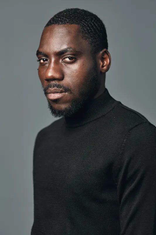 African man portrait with beard and short hair wearing black turtleneck sweater in creative conceptual visual style with serious expression on gray background.