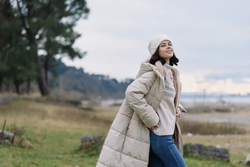 Woman in a long coat enjoying outdoor scenery in a chilly landscape, bundled in a cozy sweater and beanie, posing with a confident smile near a coastal park.