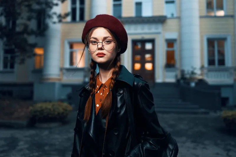 Модная женщина в красном берете и очках принимает эффектную позу на фоне очаровательного исторического кирпичного здания на оживлённой городской улице