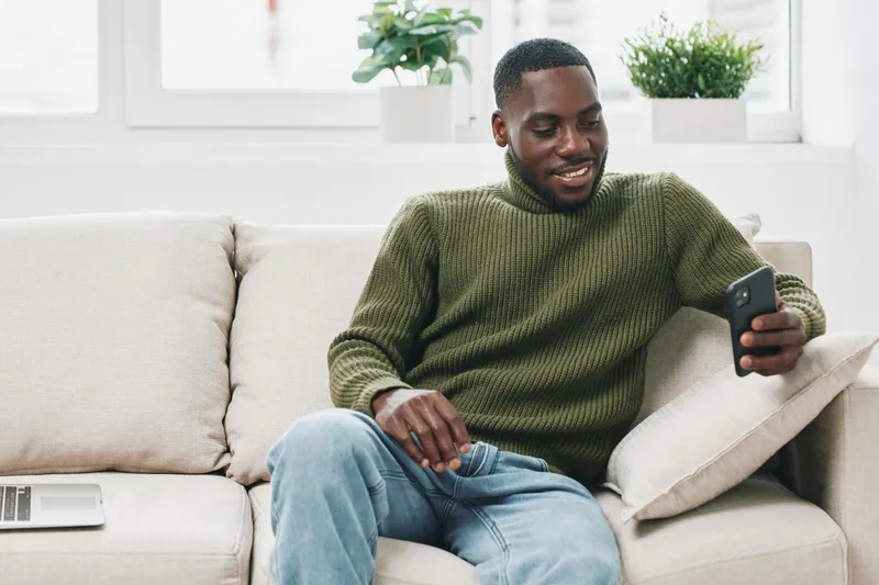 Happy African American man in green sweater enjoying smartphone on couch indoors, reflecting modern lifestyle and relaxation