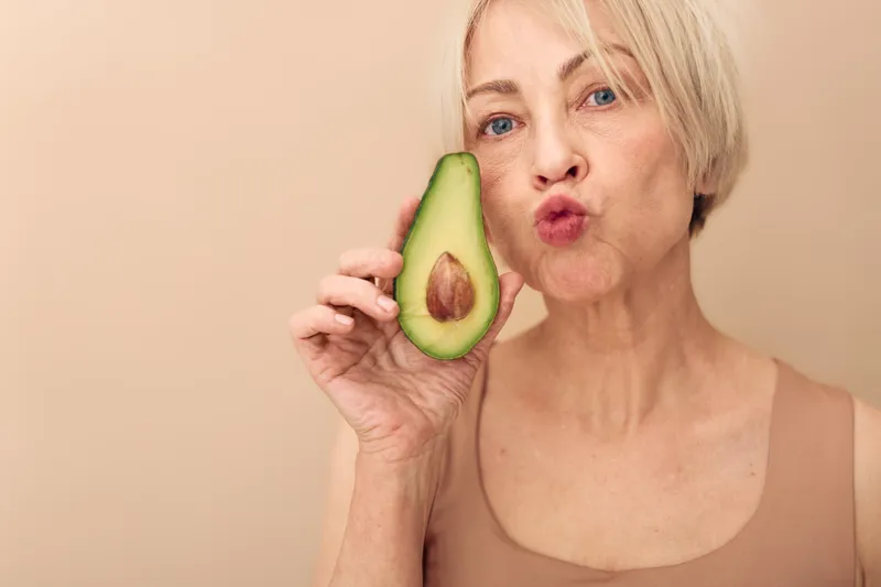 Mature woman with short blonde hair holds fresh avocado half near face, making playful kiss gesture. Concept for healthy lifestyle, nutrition, wellness, and natural beauty skin care.