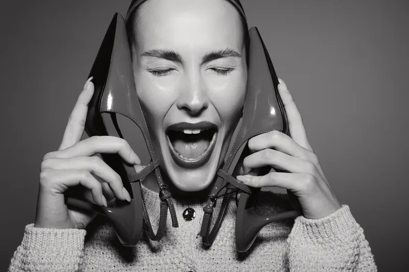 Young white woman with light skin holding two high heels near her ears, eyes closed and mouth open. Concept of stress, frustration, and emotional expression in fashion.