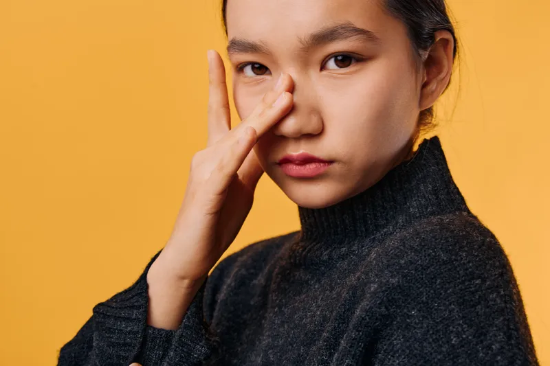 Young woman in dark sweater with pensive expression touching face against yellow background. Closeup portrait showing thoughtful mood and natural beauty with minimal makeup and hairstyle.