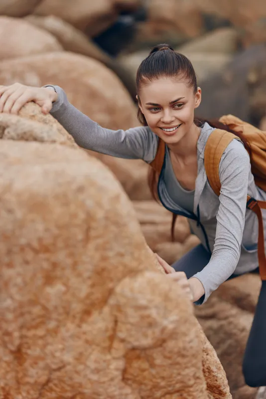 Young woman climbing rocky terrain with a backpack, showcasing determination and joy in an outdoor adventure setting, ideal for fitness and travel themes