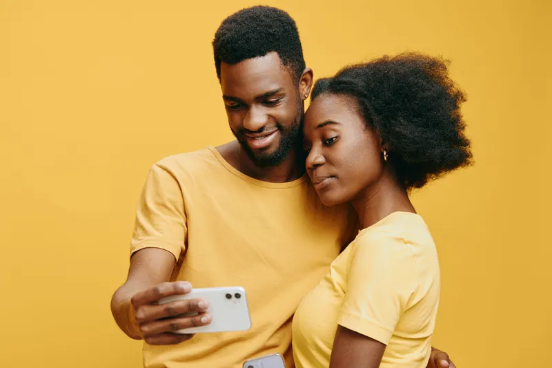 A cheerful young African couple in matching yellow t shirts smiling at a smartphone against a vibrant yellow background, embodying love, connection, and modern technology