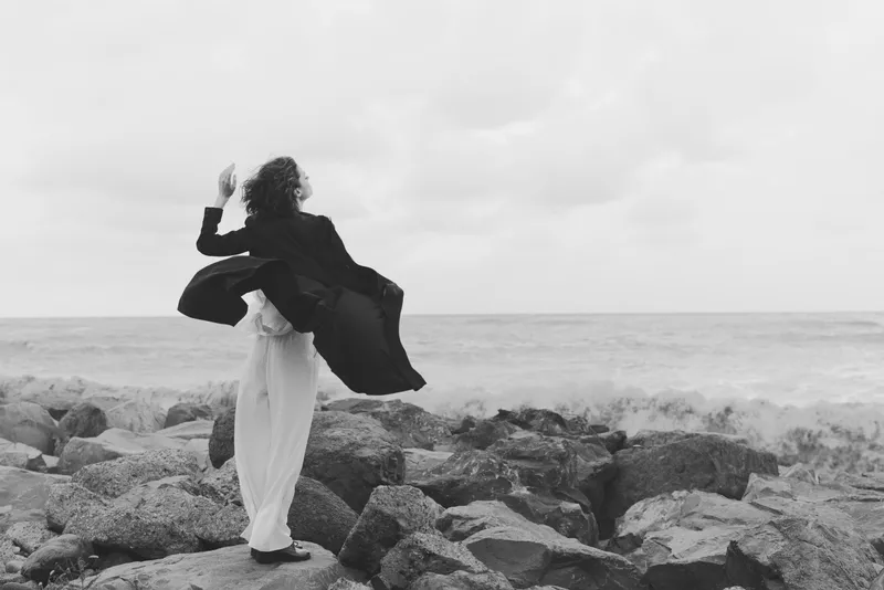 Windy coastline scene with a flowing coat over rocks by the sea, monochrome tones, a woman in motion, dramatic outdoor fashion moment, minimalist seascape, sense of freedom