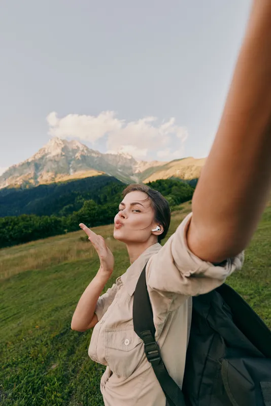 Lively woman with backpack outdoors blows a kiss toward the camera, dramatic mountain scenery behind, candid lifestyle shot, sunny day, travel inspiration, joyful moment