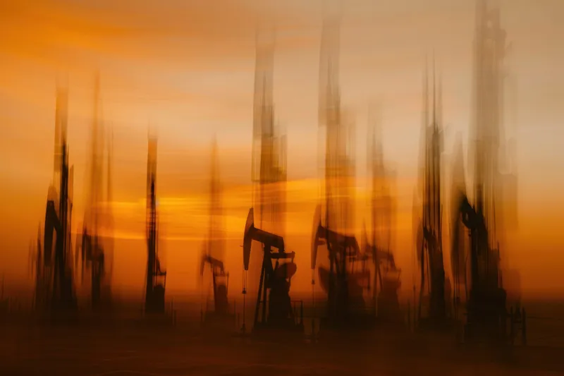 Динамичное и абстрактное изображение нефтяных вышек, силуэтами выделяющихся на фоне оранжево-золотистого неба, иллюстрирующее энергетический сектор и промышленный рост