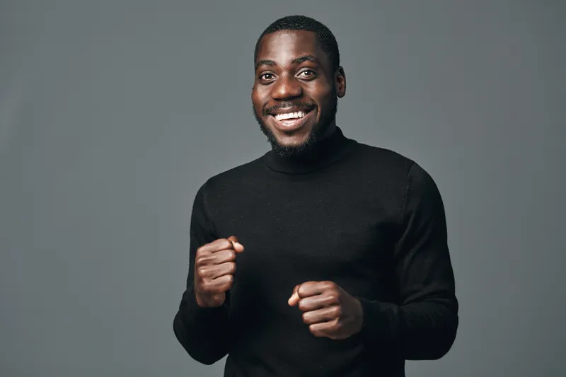 African man smiling and posing with clenched fists in creative conceptual visual style. Portrait of happy black male in black sweater with confident expression on gray background.