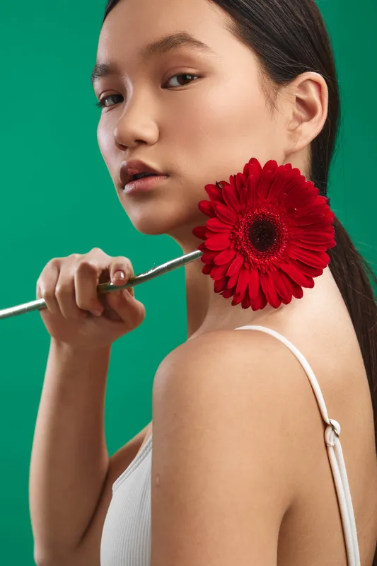 asian woman portrait with a vibrant red flower near the neck in a studio setting; calm expression, soft lighting, and a vivid green background create a stylish, elegant fashion image.