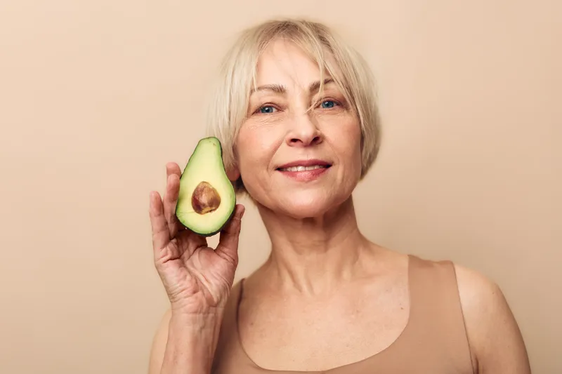 Smiling mature woman holds fresh avocado half near face on beige background. Concept of healthy lifestyle, natural beauty, skincare, wellness, nutrition, and organic food.