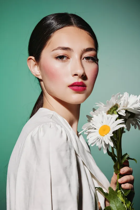 Young woman with styled hair and vibrant pink lipstick holds white daisies, posing confidently against a soft green background. Beauty concept for fashion and creative editorials.
