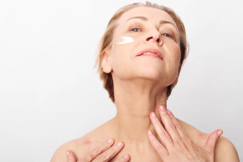 Mature woman applies white facial cream on cheek with hands touching neck. Close-up on white background. Concept of skincare, antiaging, and self-care for healthy skin and beauty.