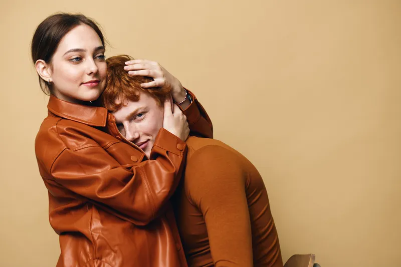 Couple in warm brown clothing hugging with affection and comfort on beige background. Young man and woman showing tender emotions and connection in casual fashion style.
