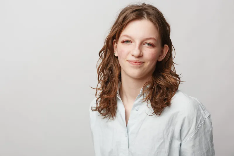 Young woman with curly brown hair smiles gently wearing a white shirt against a plain light background. Portrait of natural beauty, casual style, and confident expression.