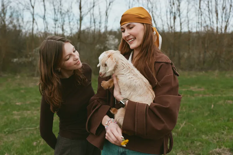 Cheerful moment outdoors with two women and a playful goat kid, warm clothing, soft sunlight, candid smiles, relaxed posture, natural colors, grass meadow background, friendly animals, joyful